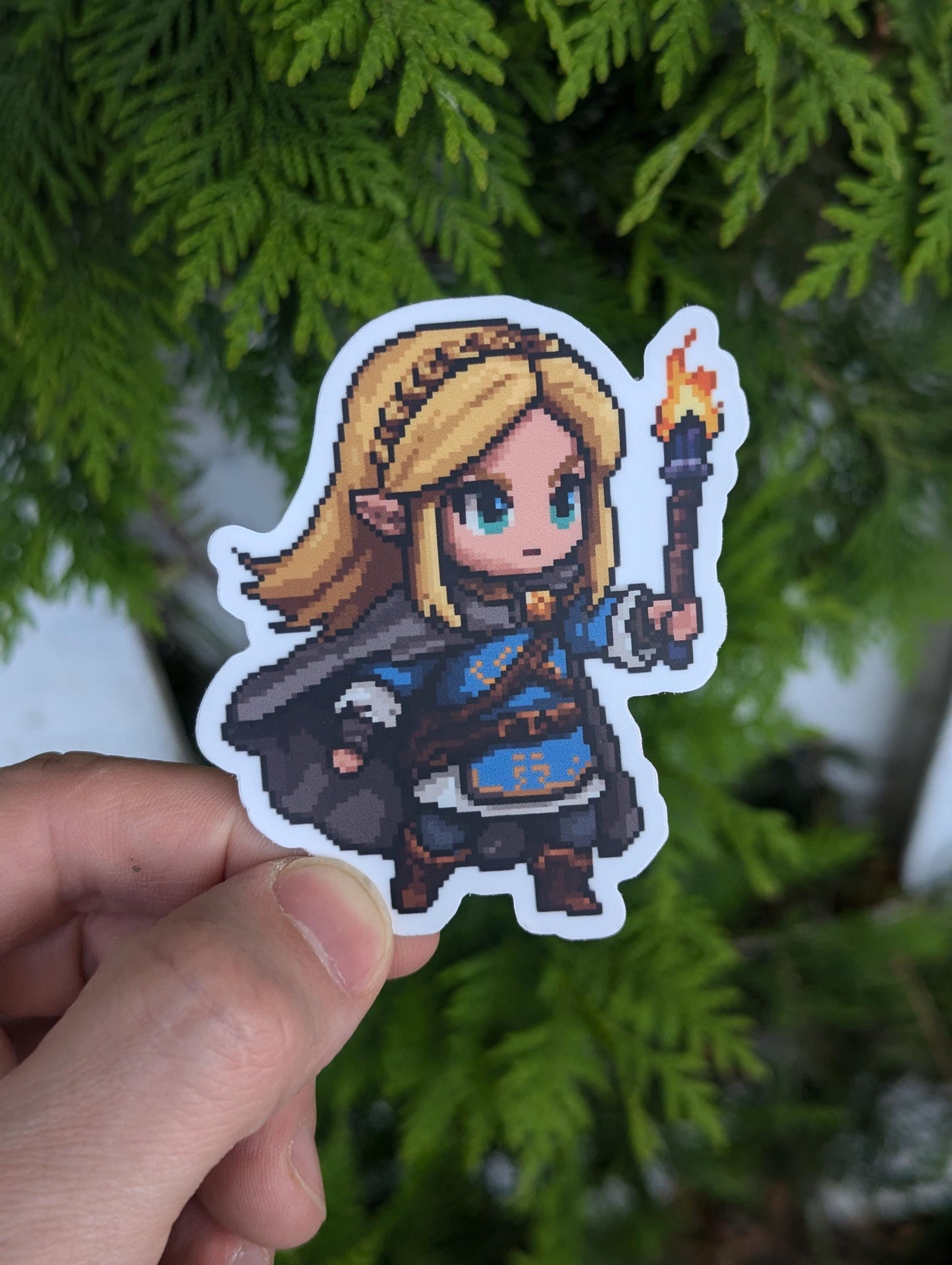 Chibi pixel-art of Princess Zelda in a blue dress and cloak, holding a lit torch aloft as her blonde hair cascades behind her.