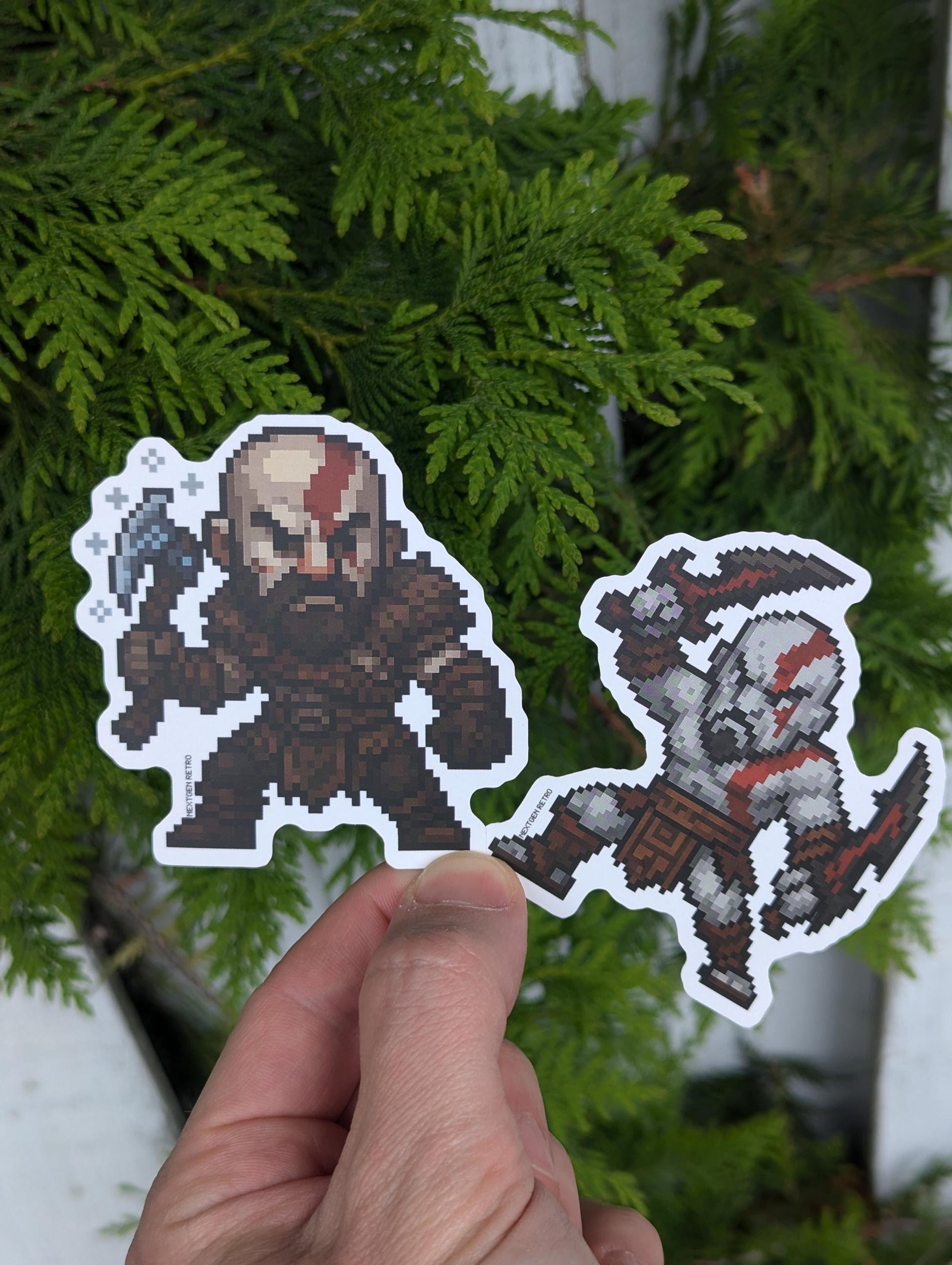 Stickers inspired by God of War. Pixelized versions of Kratos from both the Greek and Norse era games. Norse era Kratos is wearing his brown armor and holding his axe with snowflakes etched around. Greek era Kratos is pale-skinned, bare-chested and holding up the blades of chaos in both hands.
