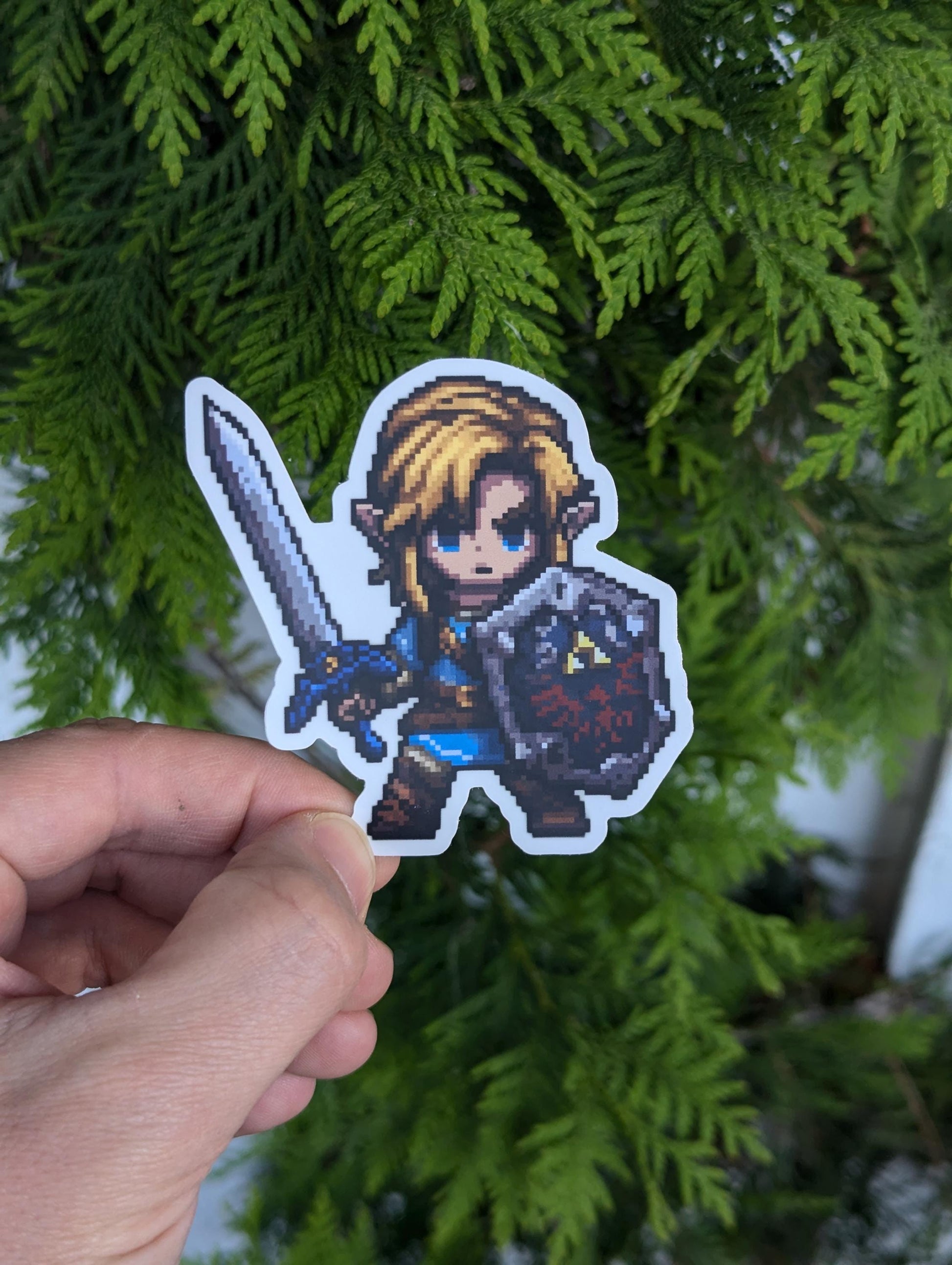 Chibi pixel-art of Link wearing a blue tunic and brown boots, gripping a sword and a Hylian shield emblazoned with the Triforce emblem.